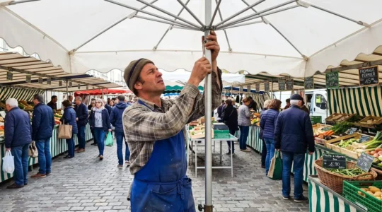 Commerçant vu de profil vérifiant le serrage du mât d'un grand parasol blanc sur un marché en plein air, geste professionnel précis, lumière naturelle matinale